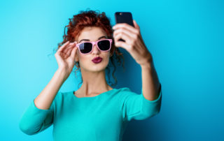 Beautiful red-haired girl in sunglasses over blue background making selfie on her cell phone. Beauty, fashion concept. Make-up and cosmetics. Studio shot.