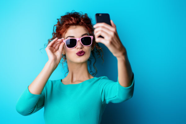 Beautiful red-haired girl in sunglasses over blue background making selfie on her cell phone. Beauty, fashion concept. Make-up and cosmetics. Studio shot.